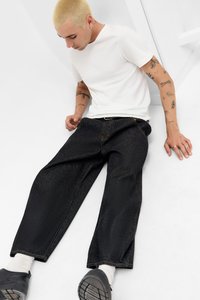 White cotton t-shirt, loose black denim pants, textured fabric, visible stitching, worn with black shoes and white socks, seated against a neutral background.