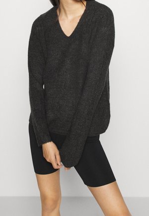 Personne portant un pull à col en V côtelé gris foncé à manches longues et un short de cycliste noir ajusté sur un fond clair uni.