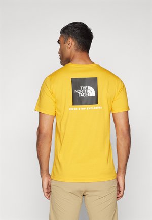 Gul kortærmet t-shirt lavet af bomuld, med et sort firkantet logo og tekst på ryggen. Enkel, afslappet design med rund halsudskæring.