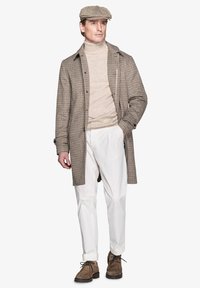 Brun houndstooth-frakke over en beige turtleneck, parret med hvide bukser og brune snørebodstøvler. Outfitet har en newsboy cap.