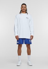 Weißes langärmeliges T-Shirt mit Logo auf der Brust, kombiniert mit blauen Mesh-Shorts, die weißen Text und ein Patch aufweisen. Weiße Socken und Turnschuhe runden den Look ab.
