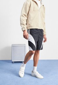 Person i beige fleece zip-up jacka, svartvit sportshorts, vita strumpor och vita kardborreskor som står på blått golv.