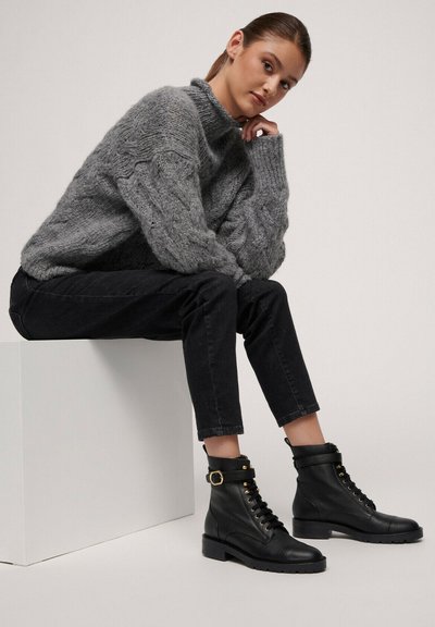 Pletena siva jopica z visokim ovratnikom, črne denim hlače in črne usnjene gležnjarje z vezanjem, okrašene z zlatimi detajli.
