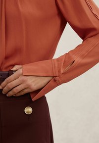 Bluza z dolgimi rokavi rjave rjave barve z naborki in z zapenjanjem na gumbe na manšetah. Oblečena preko temnordeče krila z metalnim gumbom.