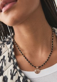 Round pendant necklace featuring black beads and gold accents, set against a patterned shirt with a quilted design.