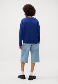 Felpa blu con vestibilità comoda, maniche lunghe e orlo a coste, abbinata a pantaloni corti in denim azzurro e calzature nere.