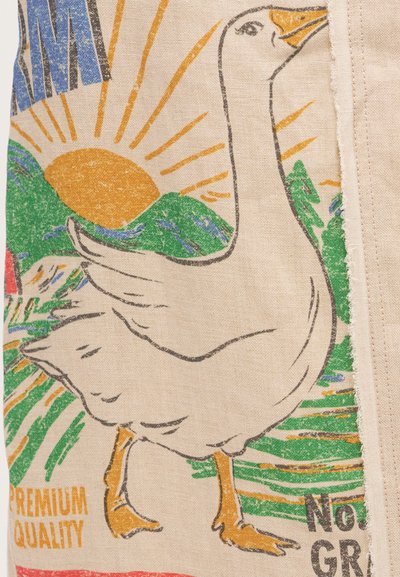 Λευκή χήνα με πορτοκαλί ράμφος και πόδια που στέκεται μπροστά από έναν ήλιο, πράσινους λόφους και κείμενο σε μπεζ ύφασμα.