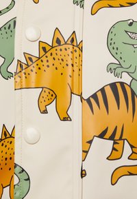 Färgglad tyg med lekfulla dinosaurieillustrationer i grönt, orange och svart på en krämfärgad bakgrund, med vita knappar.