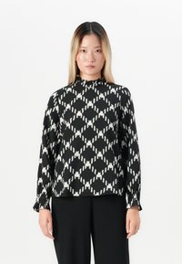 Giovane donna con capelli biondi lisci, vestita con un blusa a maniche lunghe bianca e nera a fantasia e pantaloni neri, in piedi su uno sfondo bianco.