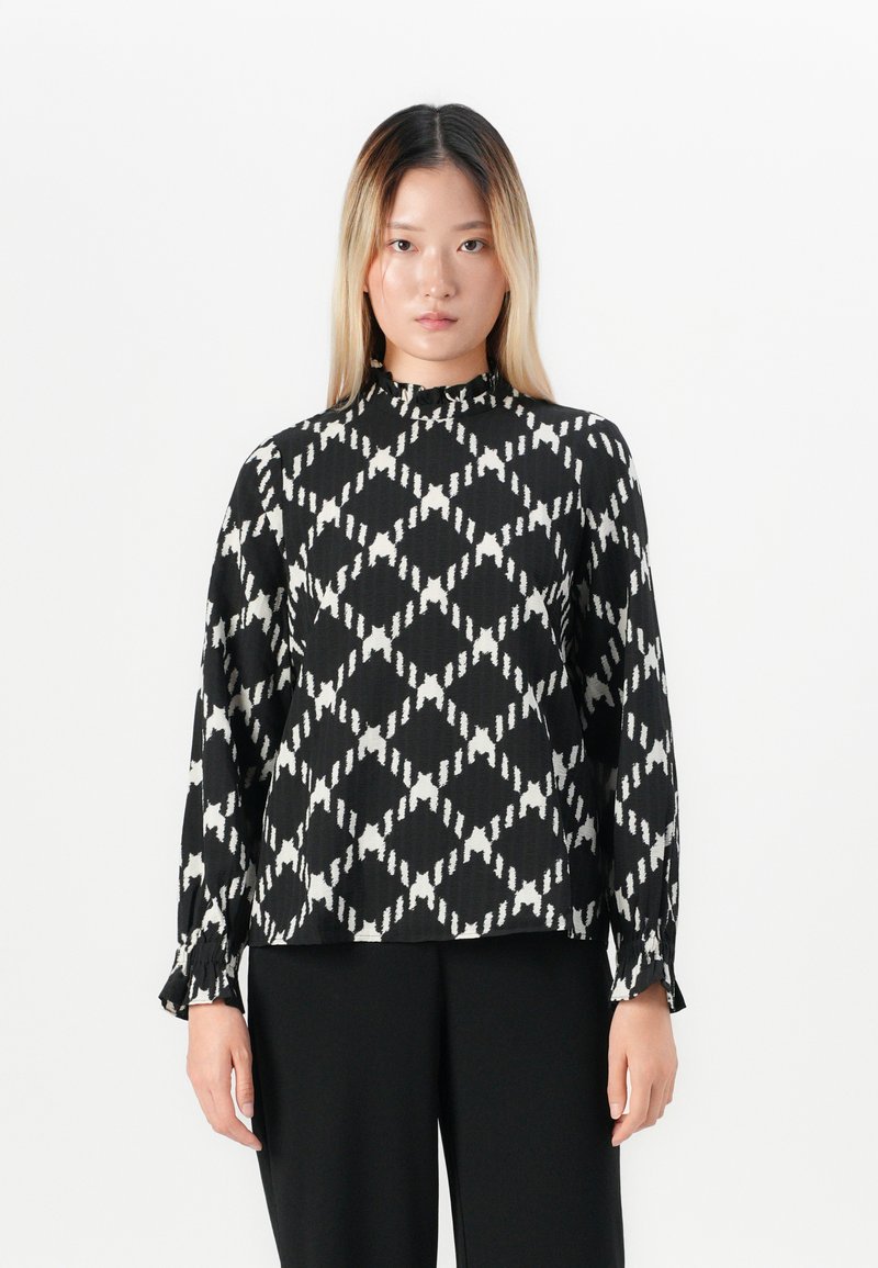 Giovane donna con capelli biondi lisci, vestita con un blusa a maniche lunghe bianca e nera a fantasia e pantaloni neri, in piedi su uno sfondo bianco.