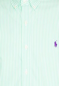 Vaaleanvihreä ja valkoinen pystyraitainen nappipaitaa, jossa on pieni violetti brodeerattu polo-pelaajan logo oikealla puolella.