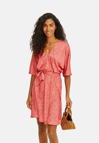 Růžové květinové šaty s výstřihem do V, krátkými rukávy a zavazováním v pase. Lehká tkanina s splývavým designem. Drží hnědou kabelku.