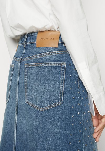 Jeansrock mit tailliertem Bund und einem hellbraunen Lederetikett, Gesäßtaschen und metallischen Nieten an einer Seite. Klassische blaue Farbe.