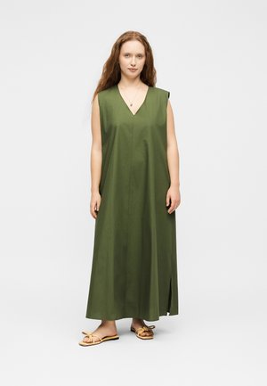 Frau mit langen roten Haaren, die ein ärmelloses olivgrünes Maxikleid und beige flache Sandalen trägt, steht vor einem einfarbigen hellen Hintergrund.