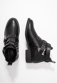 Botas negras de tobillo con tachuelas plateadas, correas con hebilla y un tacón bajo, mostradas desde ángulos superior y lateral sobre un fondo blanco.
