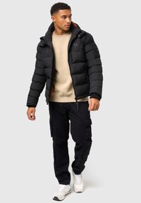 Schwarze Daunenjacke mit Kapuze, mit horizontalen Nähten und orangefarbenem Futter, getragen über einem beigen Sweatshirt und schwarzen Cargo-Hosen.