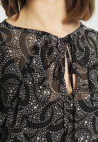 Blouse noire transparente avec motifs à pois blancs et étoiles. Elle présente une encolure en trou de serrure avec une fermeture à nouer et des bords de manches à volants.