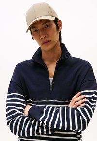 Marineblå zip-up hettegenser med hvite horisontale striper og ribbestrikkede ermer, kombinert med en lys beige caps med kontrasterende skygge.