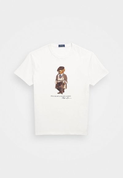 Bela bombažna majica s karikaturkastim medvedom v obleki in klobuku, z besedilom "POLO BEAR by RALPH LAUREN" in podpisom pod ilustracijo.
