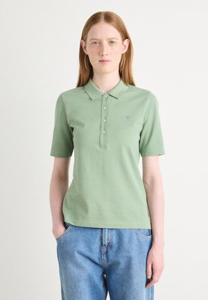 Light green polo shirt with a collar, short sleeves, and three-button placket; features a small logo on the left chest. Worn with blue jeans.