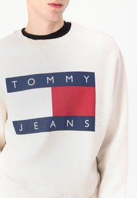 Krémová mikina s odvážnym grafickým dizajnom v námorníckej modrej a červenej farbe s textom "TOMMY" a "JEANS". Hladký lemok a manžety. Mäkkučký bavlnený materiál.