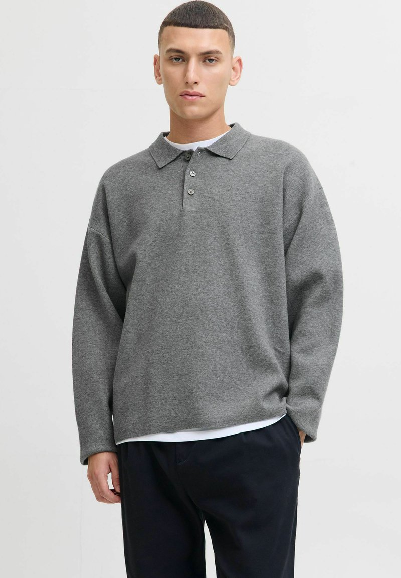 Mlađi muškarac s kratkom kosom nosi labavu svijetlosivu majicu s dugim rukavima preko bijele majice s kratkim rukavima i crnim hlačama, stoji naslonjen na bijelu pozadinu.