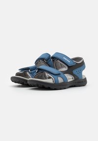 Sandalias cerradas en azul con parte superior de malla, correas de Velcro ajustables y suela de goma gris con orificios de drenaje y banda de rodadura texturizada.