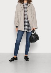 Beige Übergröße-Jacke mit zwei Fronttaschen, schwarz-graues Karomusterhemd, blaue Jeans, schwarze Lederloafers und eine gesteppt schwarze Handtasche.
