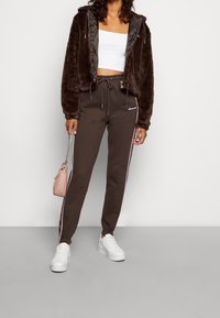 Giacca in pelliccia ecologica marrone con zip, top corto bianco, pantaloni jogger marroni con strisce rosa laterali e logo, sneakers bianche, mentre tiene una piccola borsa rosa.