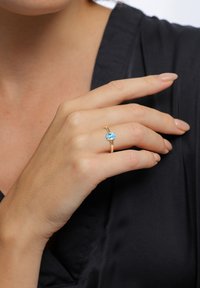 Goldring mit einem blauen ovalen Edelstein, der in einem glatten Band eingefasst ist, getragen an einer Hand vor einem schwarzen Hintergrund. Die Hautfarbe ist hell und die Nägel sind sauber manikürt.