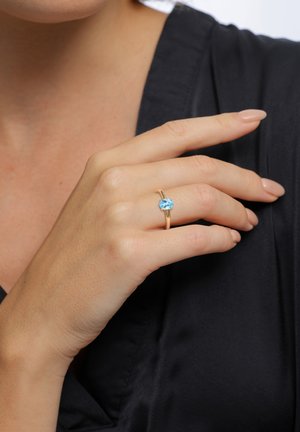 Bague en or avec une pierre précieuse ovale bleue montée sur un anneau lisse, portée sur une main contre un fond noir. Le teint de la peau est clair avec des ongles soigneusement manucurés.