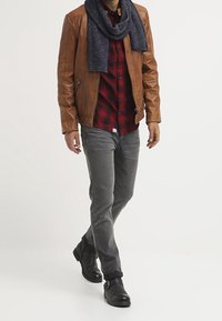 Brun læderjakke, rød ternet skjorte, grå bukser og sorte ankelstøvler. Tørklædet er mørkeblå og tilfører tekstur til outfittet.