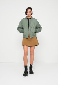 Φωτεινό πράσινο bomber μπουφάν με φερμουάρ μπροστά και ριμπ μανσέτες, συνδυασμένο με καφέ φούστα σε γραμμή Α και μαύρα μποτάκια με κορδόνια.