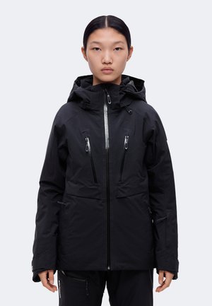 Un mannequin est de face et porte une veste noire à capuche, avec une fermeture éclair sur le devant et plusieurs poches zippées.