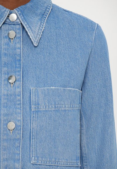 Light blue denim jacket with a button-up front, collar, single chest pocket, and silver metal buttons, showcasing a textured fabric.