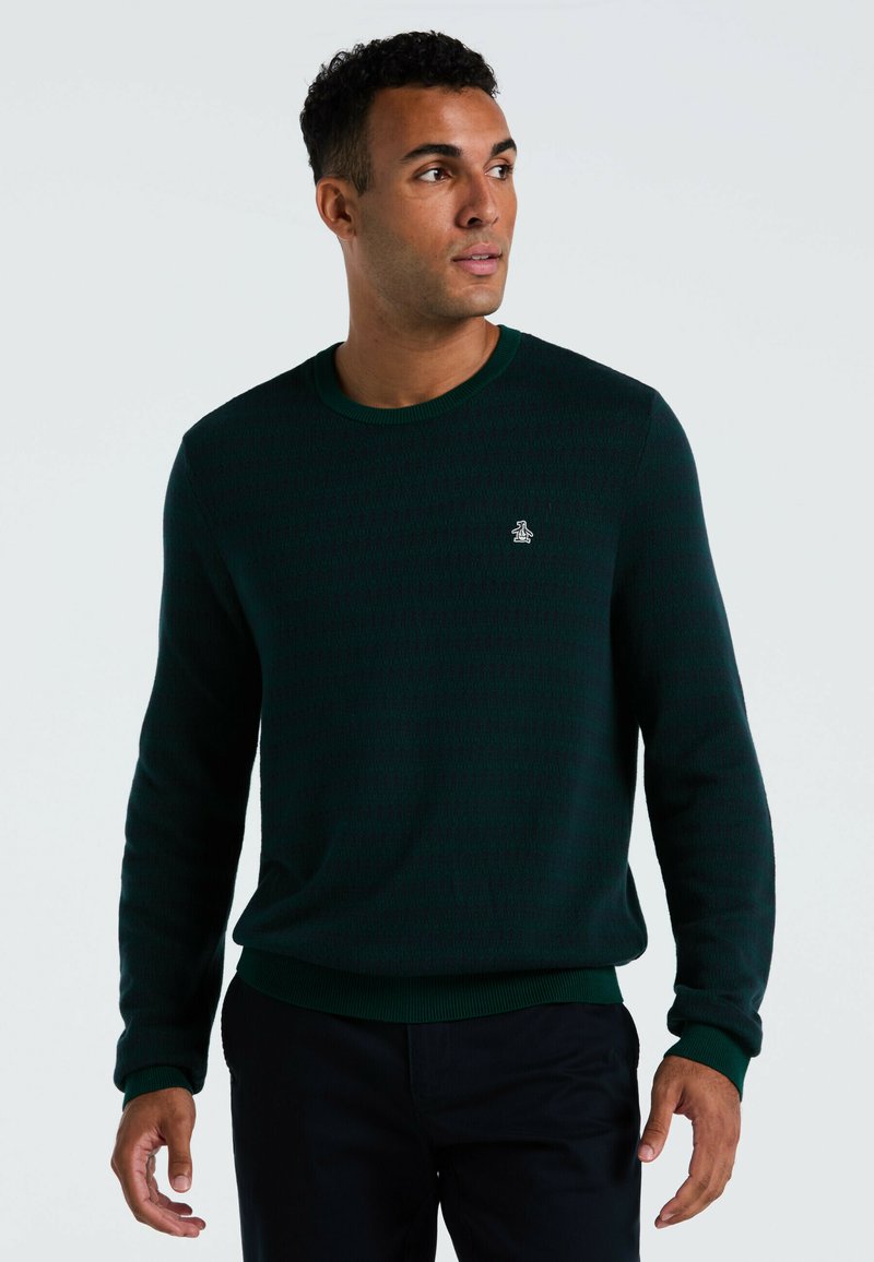 Mann, der einen dunkelgrünen Rundhals-Pullover mit einem kleinen Pinguin-Logo auf der Brust trägt, blickt nach links vor einem schlichten Hintergrund.