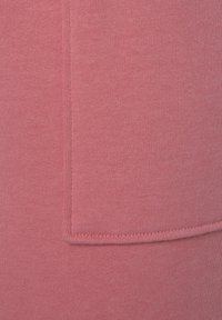 Roze gebreide stof met een gladde textuur, voorzien van een zichtbare naad en een zakdetail. Gelijke kleurverdeling over het oppervlak.