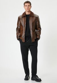 Giacca di pelle marrone con colletto in pelliccia marrone, zip frontale, due tasche a toppa, indossata sopra una camicia nera e pantaloni neri, abbinata a scarpe nere.