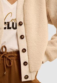 Cardigan in faux shearling color crema con orlo a coste e bottoni marroni, indossato su una t-shirt bianca con testo oro e nero, abbinato a pantaloni marroni con coulisse.