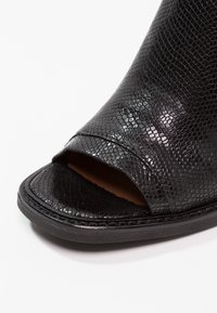 Chaussure noire à bout ouvert en cuir texturé brillant avec un motif serpent et une semelle plate sur un fond blanc.