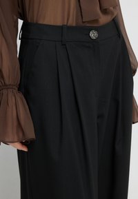 Schwarze, gefaltete Hose mit hoher Taille, ausgestattet mit einem dekorativen silbernen Knopf, und kombiniert mit einem transparenten, braunen Longsleeve-Oberteil.