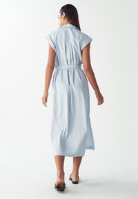 Donna che si allontana indossando un vestito midi azzurro chiaro con cintura e maniche a cappuccio, abbinato a sandali neri flat, su uno sfondo bianco.