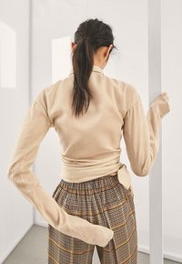 Haut beige à manches longues avec un design enveloppant, présentant des manches fluides. Associé à un pantalon à carreaux à motifs dans des tons marron, orange et crème.