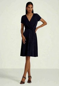 Žena stojí v krátkozrnných čiernych oblečených šatách s opaskom, doplnených čiernymi sandálmi na členkovom pásku, pózujúca pred jednoduchým pozadím.