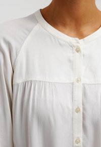 White button-up shirt with a rounded collar, raglan sleeves, and gathered texture below the yoke. Features smooth fabric and small buttons.