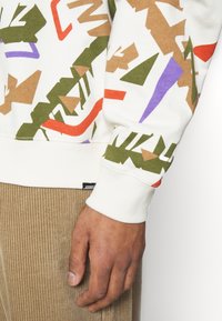 Närbild av en hand som bär en vit sweatshirt med färgglada abstrakta former och beige kordfloddsbyxor.