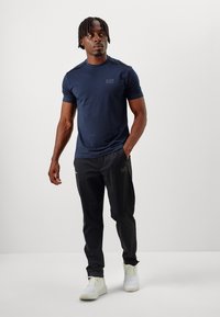 Navy cotton t-shirt with short sleeves, paired with black athletic pants. Notable EA7 logo on the shirt and pants, worn with white sneakers.