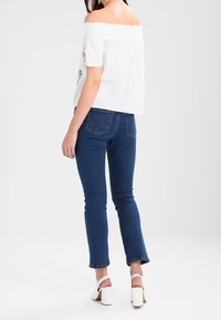 Donna con top bianco a spalle scoperte, jeans blu scuro e sandali bianchi con tacco quadrato, in piedi contro uno sfondo bianco.