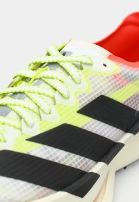 Urheilukenkä, jossa on läpikuultava päällinen verkkokuvioinnilla, neonkeltaiset nauhat, mustat raidat ja punainen sisäpinta.