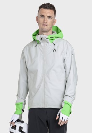 Mann trägt eine hellgraue Jacke mit neon-grüner Kapuze und Bündchen und hält einen weißen Fahrradhelm in der linken Hand.